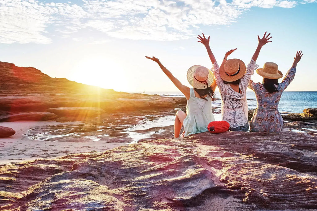 Friends raising hands at sunset on rocky beach during West Coast Road Trip. Scenic coastal moment in Western Australia.