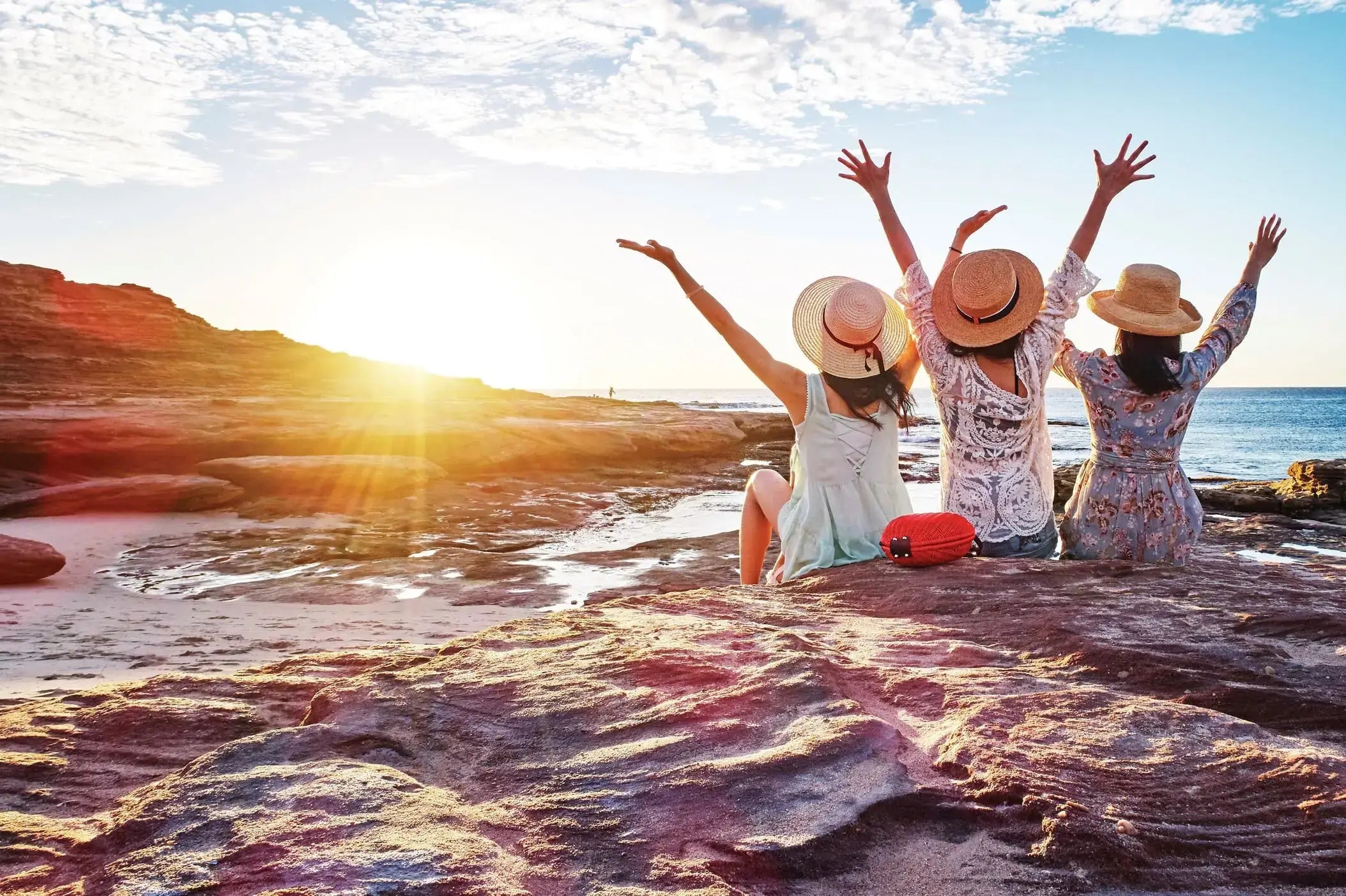 Friends raising hands at sunset on rocky beach during West Coast Road Trip. Scenic coastal moment in Western Australia.